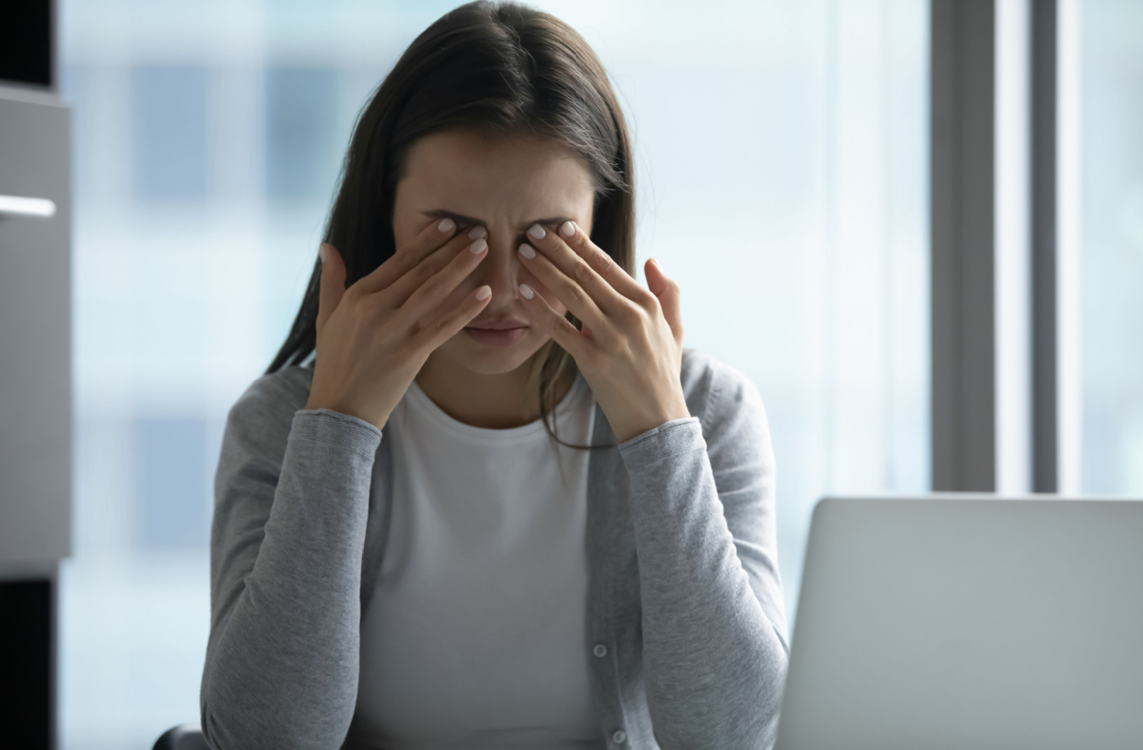 A woman with dry eyes rubs her eyes as she sits in front of a laptop.